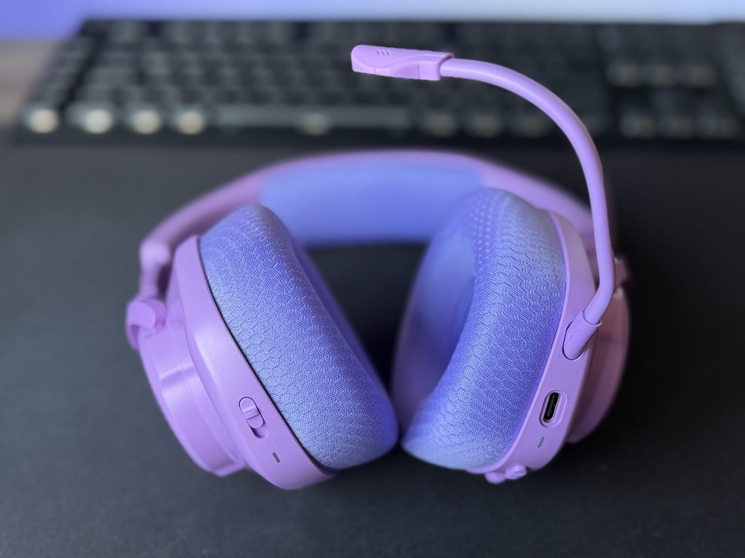 Purple JBL Quantum 360 headphones with a bendable microphone on a desk with a black mouse mat beneath and a black keyboard in the background.