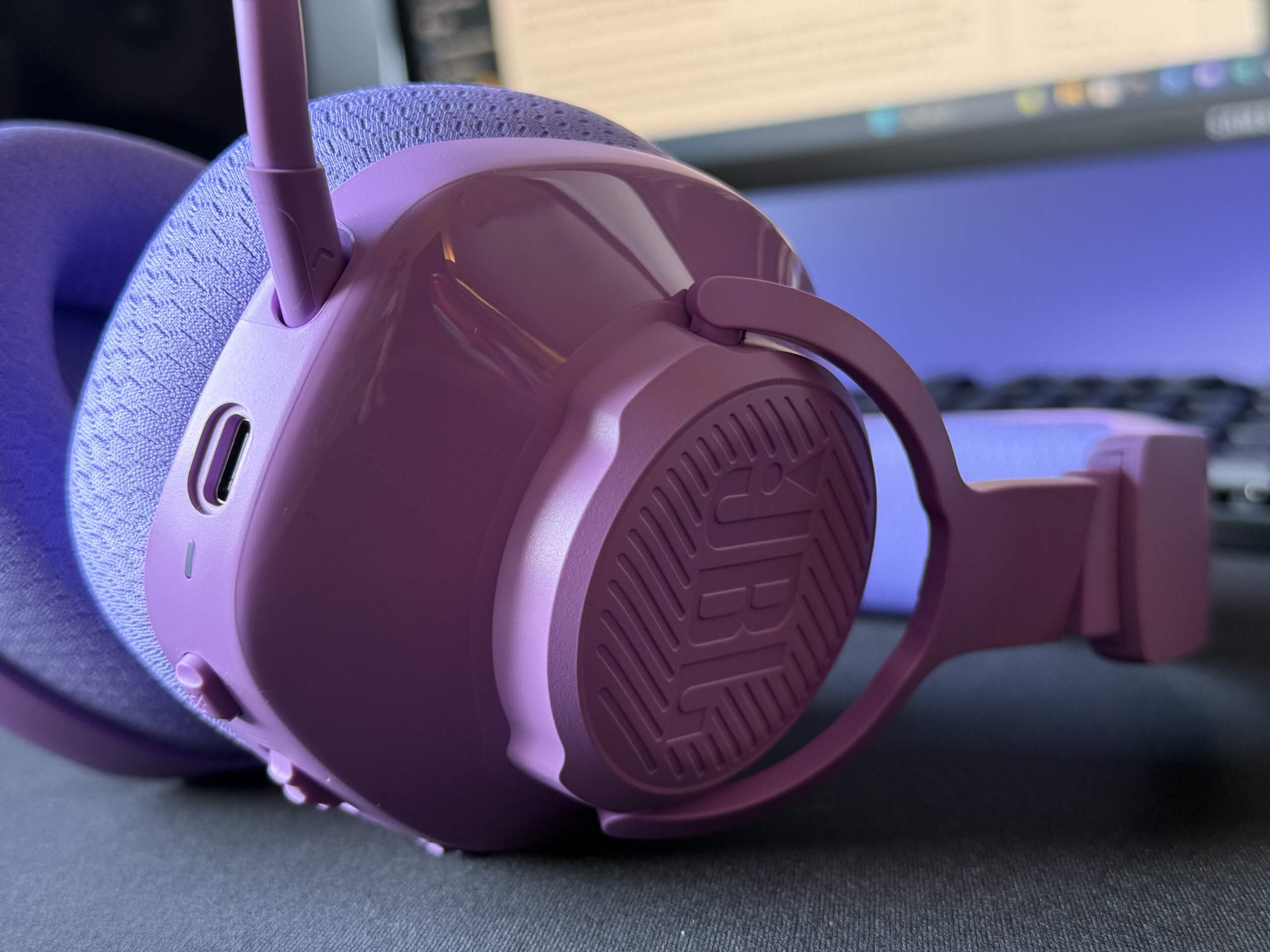 Close-up of the side of the JBL Quantum 360 headset in purple over-ear headphone with a textured design on a blurred keyboard and monitor background.