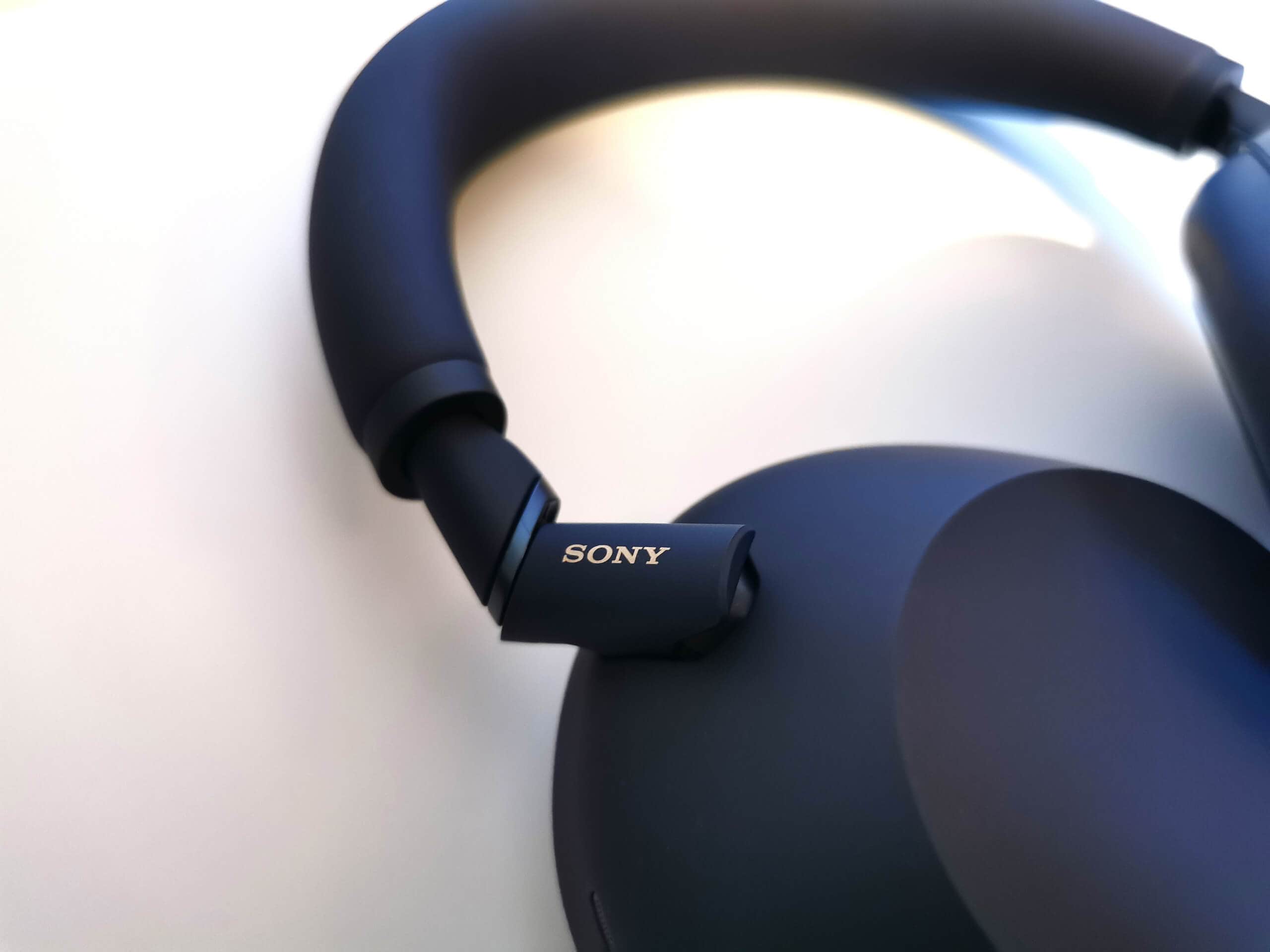 Close up of the Midnight Blue Sony WH-1000XM6 brand new metal swivel hinges. With a gold Sony logo on the side. Over a blurred white background.
