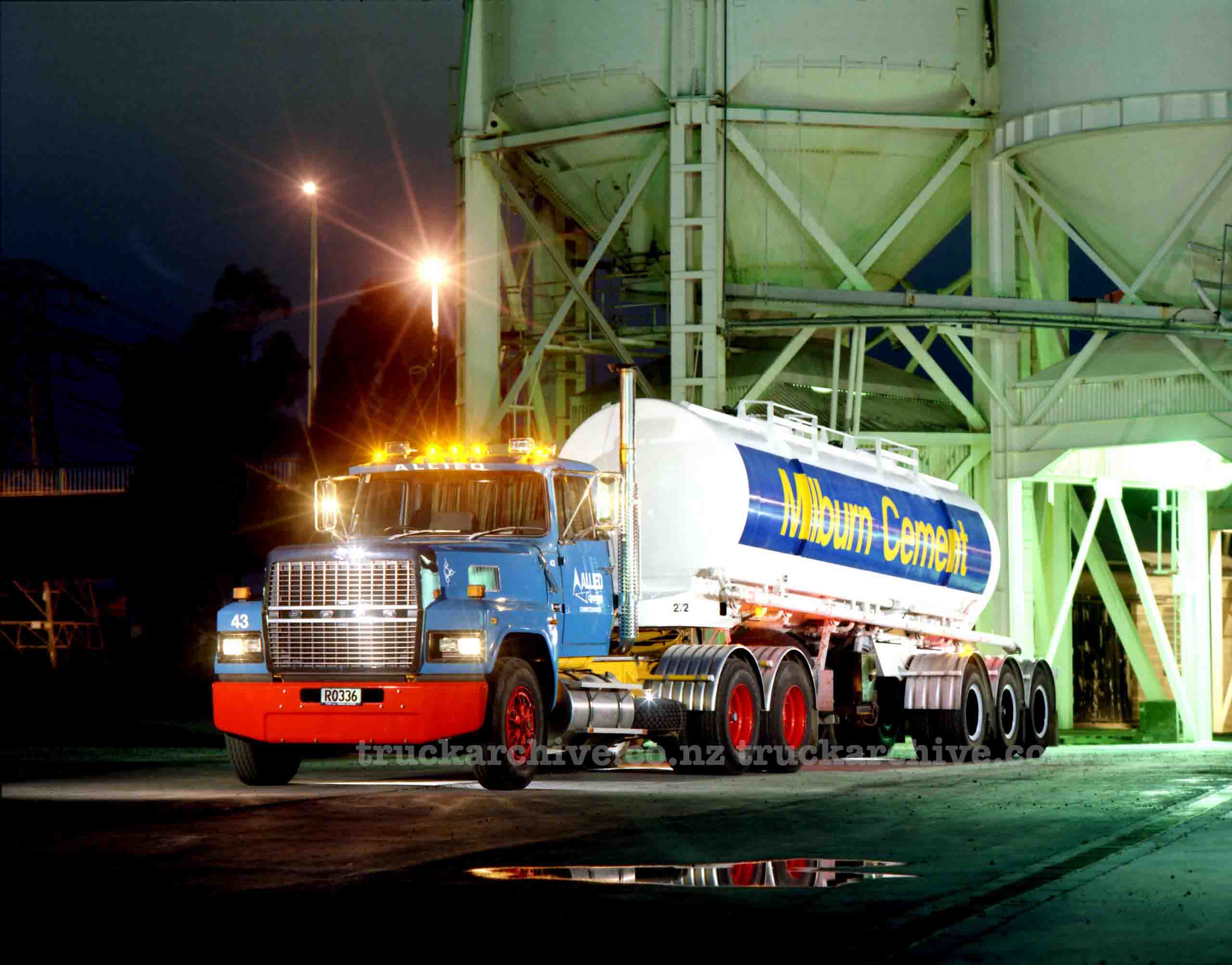 Fuel tanker truck parked at night near industrial silos, illuminated by nearby lights.