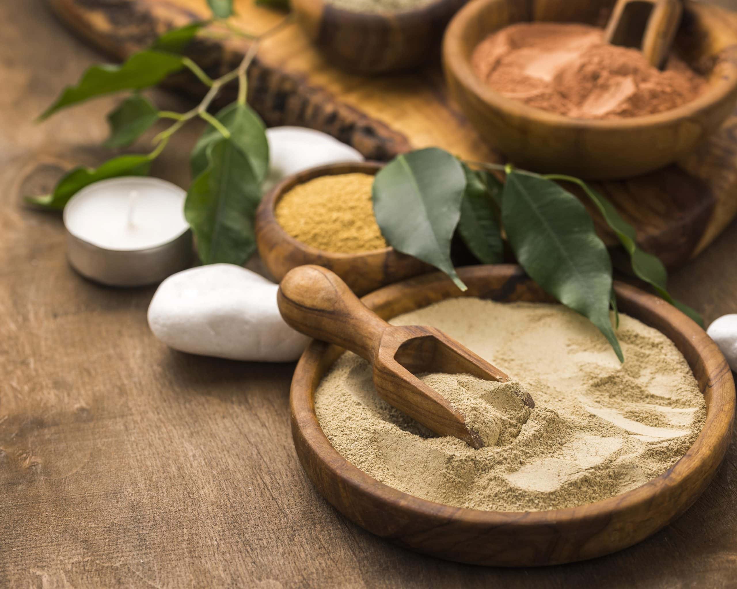 Udhwarthanam - An overhead, close up shot features several wooden bowls filled with various shades of cosmetic clay powder, including beige, ochre, and terracotta. In the foreground, a large wooden bowl contains light beige clay with a small wooden scoop resting inside. The scene is arranged on a rustic wooden surface and accented with smooth white stones, green leaves, and a small white tealight candle, evoking a natural, spa like atmosphere.