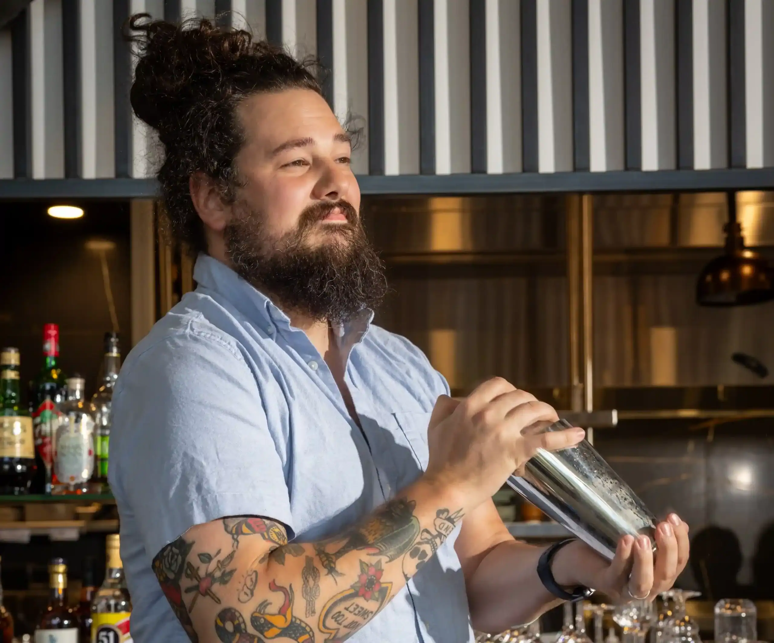 Professional bartender shaking a cocktail in a modern Raleigh bar with premium spirits, bar tools, and an open kitchen in the background.