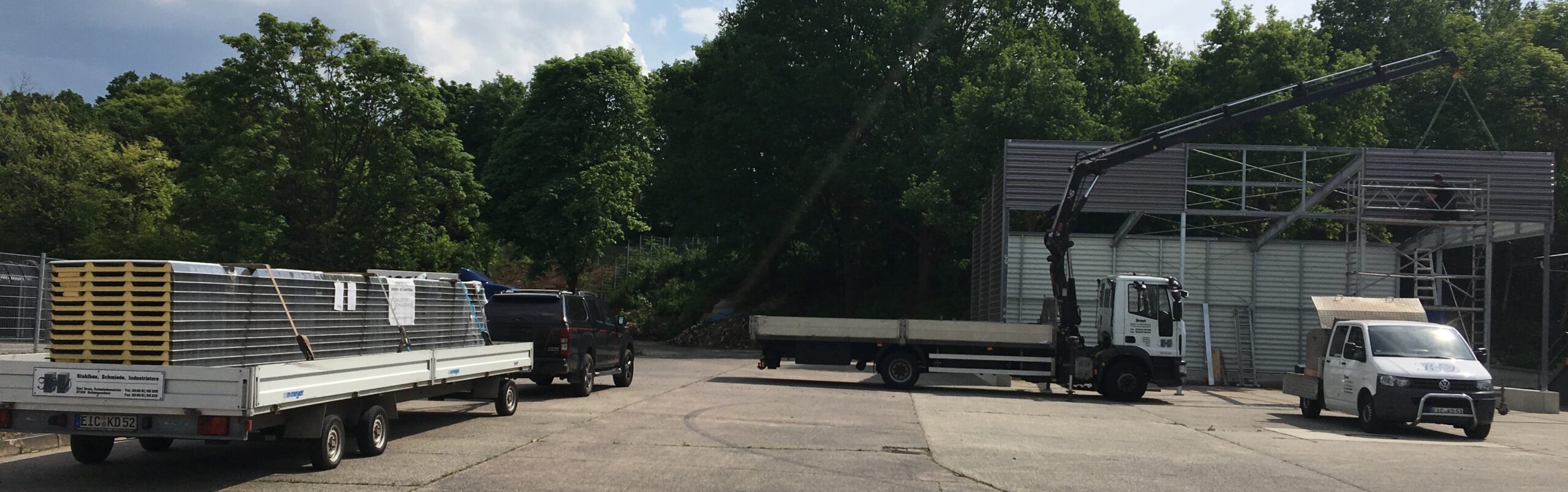 Baustelle von Stahl- und Metallbau Drost: Kran-LKW montiert eine Stahlkonstruktion, daneben Materiallieferung mit Sandwichpaneelen auf Anhänger.