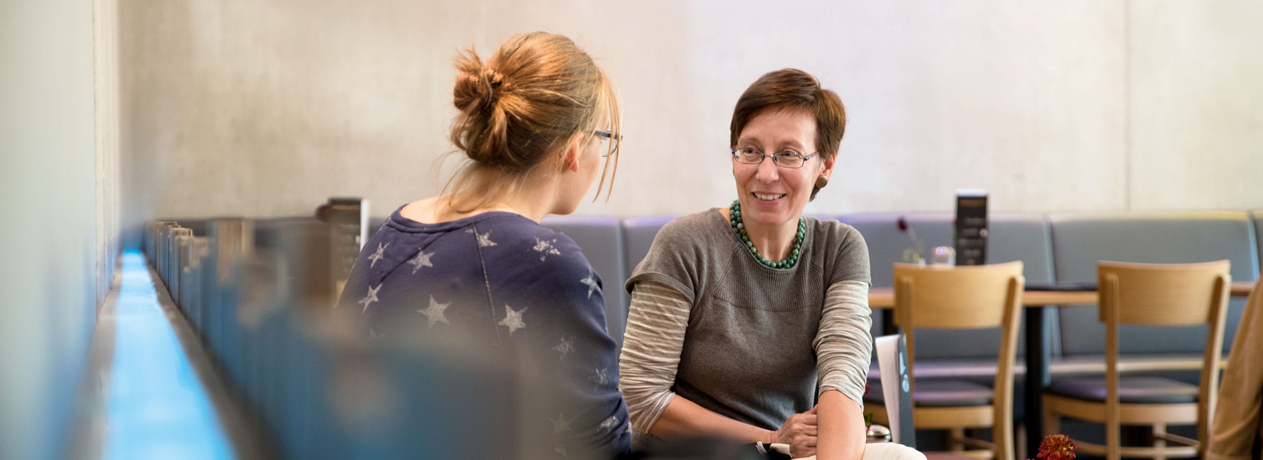 Zwei Frauen sitzen einander zugewandt in einem Raum und unterhalten sich. Eine trägt einen dunkelblauen Pullover mit Sternenmuster, die andere lächelt freundlich und trägt eine Brille sowie eine Kette. Im Hintergrund stehen Tische und Stühle.