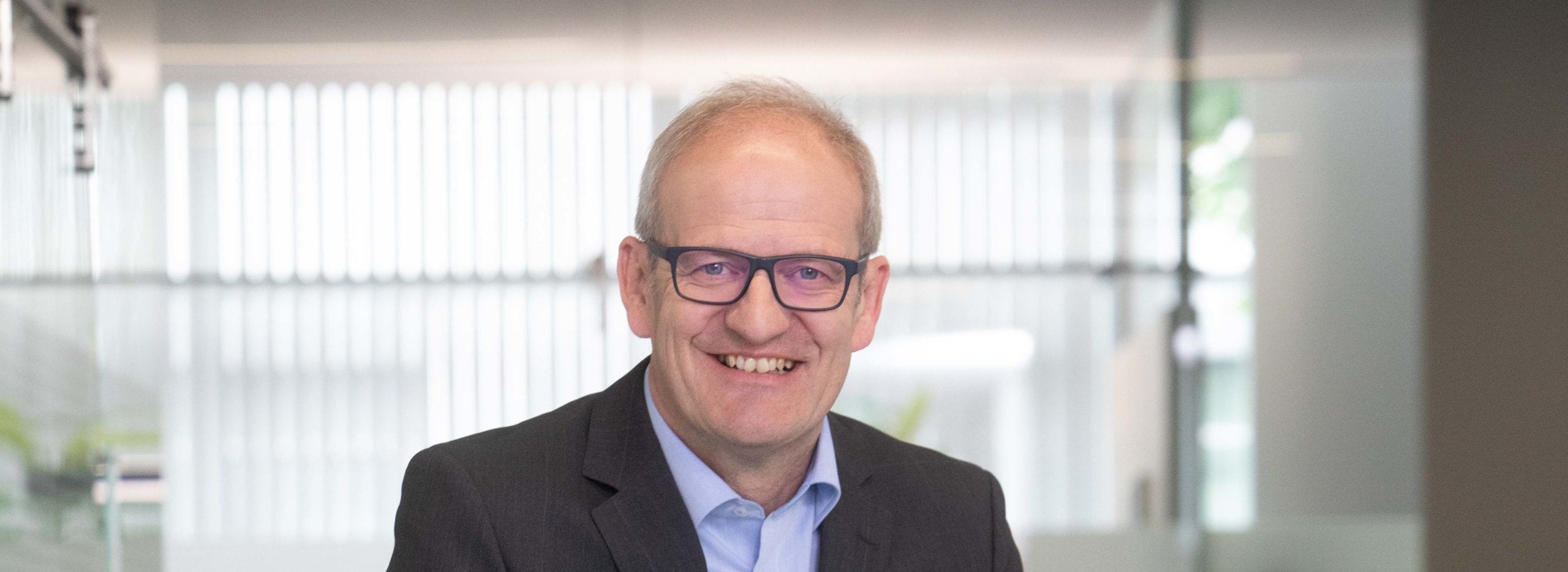 Ein älterer Mann mit kurzem hellgrauem Haar, Brille und Anzug lächelt freundlich in einer hellen, modernen Büroumgebung.