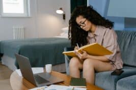A girl sees a Clinical Psychologist remotely on her laptop and takes notes in a journal, while sitting on a couch