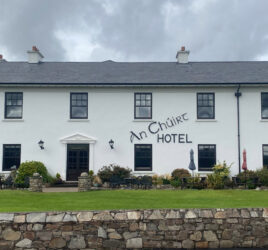 White two-story building with "An Chuirt Hotel" sign, a green lawn, stone wall, and cloudy sky in the background.