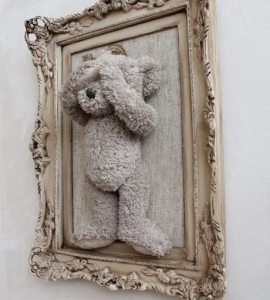 Soft gray teddy bear plush toy framed in an ornate vintage-style frame, creating a cozy and stylish wall decor piece. Perfect for adding a touch of comfort and charm to any living space.