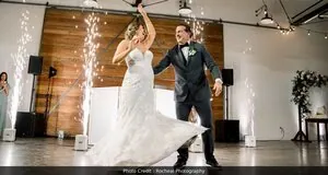 This image highlights a smiling bride and groom during their first dance. The groom is spinning his bride around. There are a set of large doors in the back drop, as well as a DJ setup. Cold sparklers are shinning in the background, illuminating the scene and providing a breathtaking atmosphere.