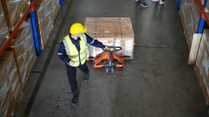 Warehouse worker using a pallet jack with a pallet full of carboard boxes.