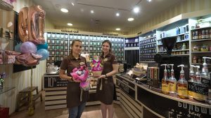Две молодые девушки-продавца с собранными волосами и открытыми улыбками стоят в полный рост за прилавком кофейно-чайного магазина. Каждая из них держит в руках празднично упакованный подарочный набор. Обе одеты в одинаковую фирменную форму: коричневые футболки-поло и темные фартуки с логотипом. На заднем плане виден темный деревянный стеллаж со множеством банок для развесного чая с надписями «ЧЕРНЫЙ ЧАЙ» и «ЗЕЛЕНЫЙ ЧАЙ». Справа на переднем плане расположена барная стойка с кофемолкой, сиропами и надписью «БОЛЬШОЙ выбор». Слева видны надувные фольгированные шары.