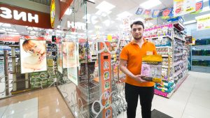 Молодой парень с короткой бородой и открытой улыбкой стоит в полный рост в проходе зоомагазина. В руках он держит и демонстрирует большую желтую упаковку сухого корма для животных бренда «GRANDIN». На нем ярко-оранжевая футболка-поло и черные брюки. Вокруг него расположены стеллажи, плотно заставленные товарами для животных, кормами, наполнителями и аксессуарами. На заднем плане видна стеклянная входная группа магазина.
