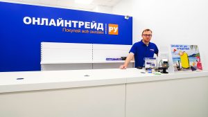 Мужчина среднего возраста в очках с короткой стрижкой и сдержанной полуулыбкой стоит за белой длинной стойкой выдачи заказов. Он запечатлен по пояс, слегка опирается одной рукой на прилавок, на котором установлен платежный терминал. Одет в ярко-синюю футболку-поло. Позади него — большая синяя стена с фирменным логотипом «ОНЛАЙНТРЕЙД РУ Покупай всё онлайн».