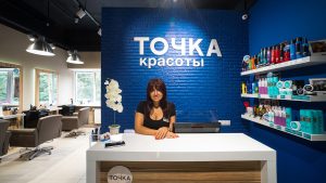Молодая женщина с темными длинными волосами, челкой и открытой улыбкой сидит за белой стойкой администратора в салоне красоты. Она находится в открытой позе, сложив руки перед собой на столе. Одета в черную футболку. Позади нее — ярко-синяя кирпичная стена с крупной белой вывеской «ТОЧКА красоты» и полками с косметическими средствами для волос. Слева виден парикмахерский зал с креслами и зеркалами.