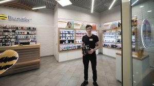 Молодой парень с короткой стрижкой и открытой улыбкой стоит в полный рост в центре салона связи. Он держит в руках и демонстрирует товар — черную коробку со смартфоном или аксессуаром. На нем черная футболка с фирменным бейджем и черные брюки с кроссовками. Слева видна брендированная деревянная стойка с логотипом «Билайн», на заднем плане — белые витрины со смартфонами, гаджетами и аксессуарами.