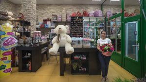 Молодая девушка с собранными в хвост волосами и сдержанной полуулыбкой стоит в полный рост в цветочном магазине. В руках она держит и демонстрирует круглый букет из розовых роз. На ней белая футболка с коротким рукавом и темные джинсы. Слева от нее расположен коричневый деревянный прилавок, на котором сидит большой плюшевый медведь кремового цвета. На полках позади видны подарочные коробки, воздушные шары и упаковочные материалы. Справа находится зеленая пластиковая входная группа со стеклянной дверью.