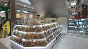 Молодой парень восточной внешности с короткой стрижкой и открытой улыбкой стоит за большим угловым торговым островком с сухофруктами и орехами. Он запечатлен по пояс, приветственно подняв одну руку. Одет в светлую футболку. Витрины островка со светодиодной подсветкой доверху заполнены разнообразными орехами, цукатами и сухофруктами, разложенными по ячейкам. На заднем плане виден интерьер коридора ТРЦ, павильон «Связной» и вывеска «РЕМОНТ СОТОВЫХ».