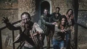Grupo de personas disfrazadas de zombis, con ropa desgastada y aspecto aterrador, caminando sobre una pasarela de metal.
