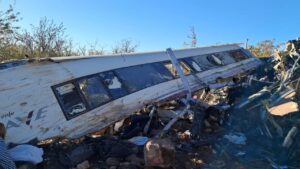 Un tren volcado y dañado en un área desolada, rodeado de escombros y vegetación. Se observan partes del autobús y algunos