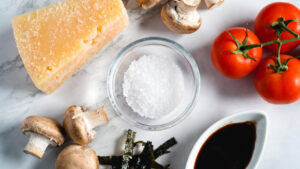 Bowl of MSG crystals surrounded by naturally glutamate-rich foods including parmesan, tomatoes, mushrooms, soy sauce, and kombu seaweed