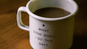 A white mug with black text that reads, "WHAT GOOD SHALL I DO THIS DAY?" sits on a wooden surface, filled with coffee or tea, inspiring thoughts of successful career networking.