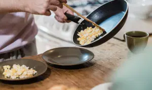 Persona sirviendo huevos revueltos recién preparados desde un sartén negro hacia un plato en una mesa con pan y café.