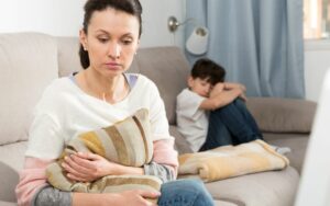 Ich kann mein Kind nicht mehr ertragen, Frau sitzt mit Kissen auf dem Sofa, macht sich sorgen, Kind dahinter zusammengekauert, traurig