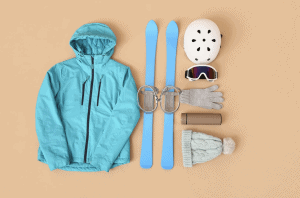 Flat lay of ski trip packing list essentials including a teal ski jacket, blue skis with bindings, white helmet, ski goggles, gloves, knit hat, and thermos on a neutral background.