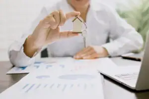 man holding a miniature house with keys, sitting at a desk with graphs and papers on it