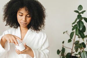 A black woman applies skincare cream indoors, wearing a white robe beside a green plant.