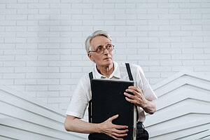 Elderly man in glasses and suspenders holding a briefcase in a modern office setting.