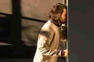 Man with shoulder-length hair in a beige jacket leans against a doorway, representing the need for therapy for men in Detroit and support from a therapist for men in Michigan.