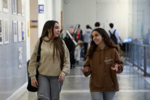 Dos jóvenes conversan en un pasillo universitario, rodeadas de estudiantes y obras en las paredes, en Alcalá de Henares.