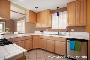 Bright kitchen at 2416 Vancouver Ave in North Park featuring light wood cabinetry, white tile countertops, a stainless steel dishwasher, and a breakfast bar overlooking the dining area.