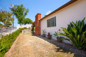 Side yard at 3727 Suffolk Drive in Rolando Park featuring a prominent red brick chimney, manicured green hedge, and potted tropical plants along the side of the home.