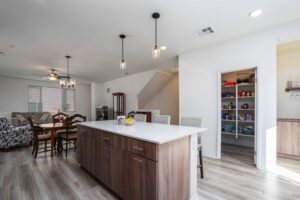 Urban-inspired modern kitchen at 5235 Beachfront Cove Unit 102 showcasing a spacious white quartz island, wood-grain cabinetry, walk-in pantry, and view to the open dining and living room area