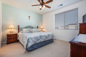 Modern bedroom at 5235 Beachfront Cove Unit 102 featuring a light blue accent wall, wooden ceiling fan, and custom vinyl blinds in the Playa Del Sol gated community.