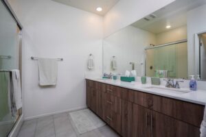 Modern primary ensuite bathroom at 5235 Beachfront Cove Unit 102 in Playa Del Sol, featuring a spacious double vanity with wood-finish cabinets, white stone countertops, and a large custom mirror reflecting a glass-enclosed walk-in shower.