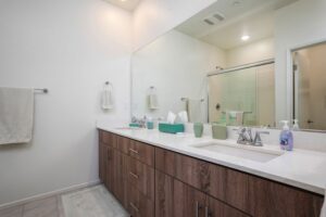 Modern primary ensuite bathroom at 5235 Beachfront Cove Unit 102 in Playa Del Sol, featuring a spacious double vanity with wood-finish cabinets, white stone countertops, and a large custom mirror reflecting a glass-enclosed walk-in shower.