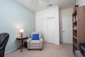 Cozy guest bedroom or home office at 5235 Beachfront Cove Unit 102 featuring a plaid armchair, wooden bookshelf, and neutral carpet.