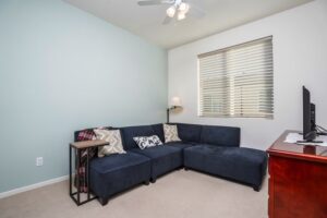 Private bedroom at 5235 Beachfront Cove Unit 102 featuring a blue accent wall, navy sectional sofa, ceiling fan, and window with custom vinyl blinds.
