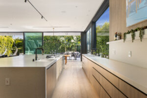 Contemporary kitchen with minimalist design, white surfaces, natural wood cabinetry, expansive glass sliding doors showing lush greenery outside.