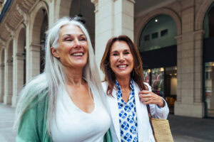 Two women, one with long gray hair and a green cardigan, and the other with brown hair and a white blazer, smile while standing outside a building with arched architecture, celebrating a successful mid-career transition.