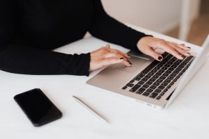 A woman typing on a laptop with a cell phone.