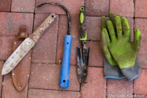 Essential gardening tools including hori hori knife, cobra head weeder, trowel, and green gloves.