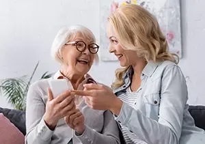 Smiling elderly woman receiving friendly home care support as caregiver helps her use a smartphone.