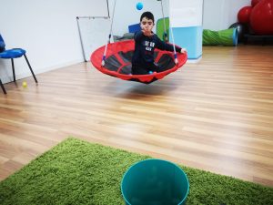 sala_terapia_ocupacional_centro_neures_barcelona_niño_en_columpio_lanza_pelota_a_encestar_en_un_cubo