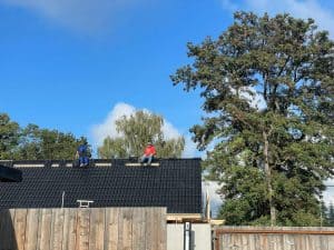 Dachdecker bei der Arbeit auf dem Dach eines Hauses, umgeben von Bäumen und einem klaren blauen Himmel.