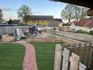 Neuer Garten mit Baustelle, grüne Rasenfläche, rot-steinpfad, Holzzäune, im Hintergrund alte und neue Gebäuden, im sonnigen Himmel, Natur.
