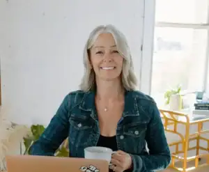 Hafdis holding a mug and sitting on a desk in front of a laptop smiling at the camera